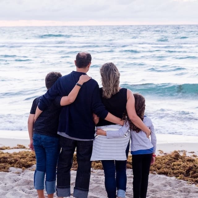 Family of four stands on the beach in eachothers arms.